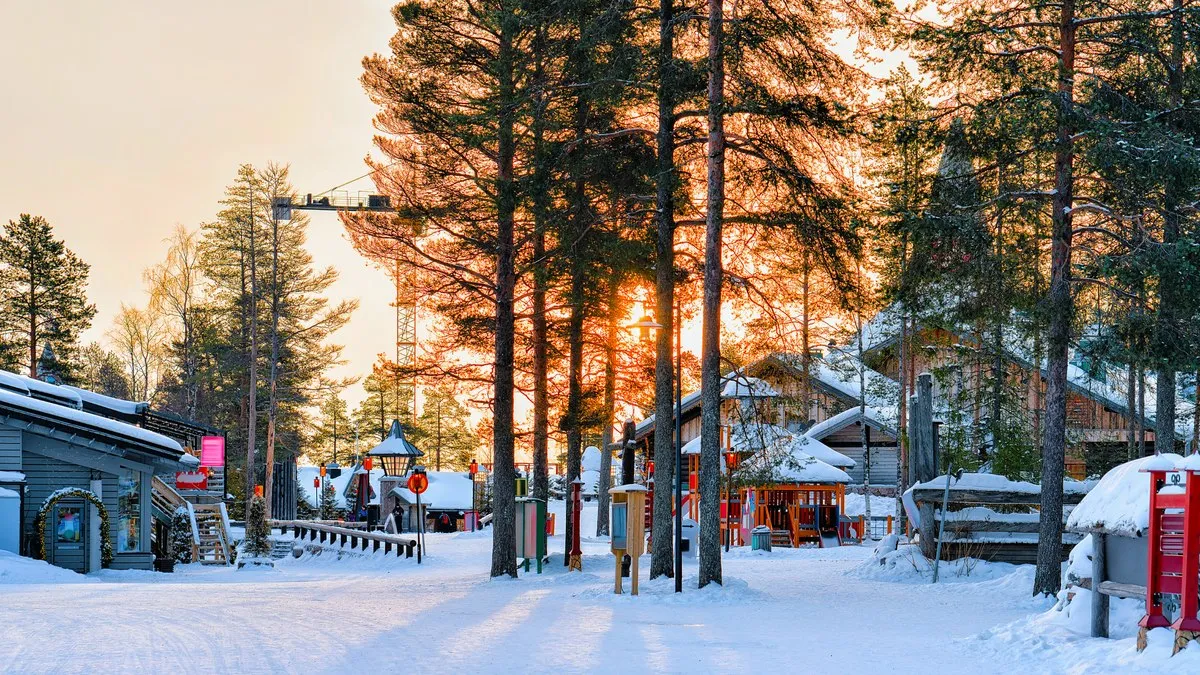 Rovaniemi’deki Noel Baba Köyü’nde karla kaplı ahşap evler ve çam ağaçları arasından batan güneş