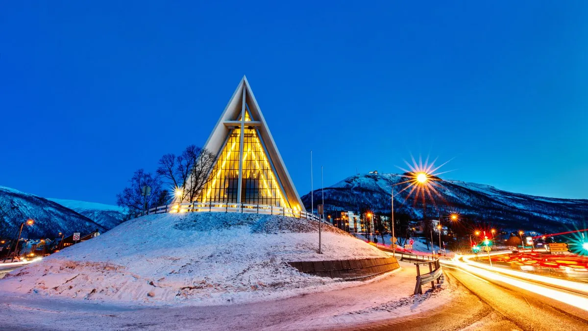 Tromsø’daki Arktik Katedrali’nin karla kaplı tepe üzerinde ışıklandırılmış üçgen cephesi ve arkadaki karlı dağ