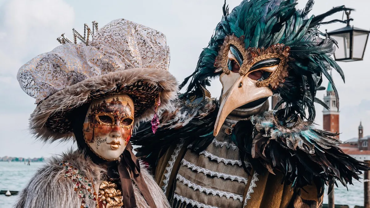 Venedik Karnavalı’nda tüylü kostümler ve geleneksel maskelerle poz veren iki kişi, arkada San Giorgio Maggiore çan kulesi